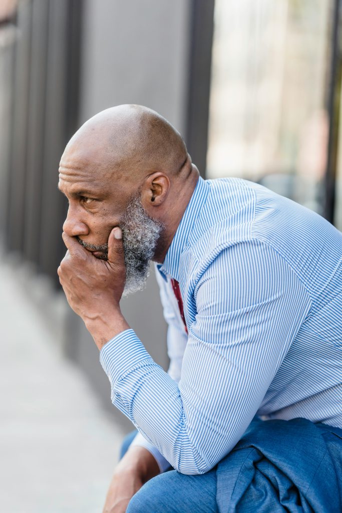 Side view of frustrated bearded African American businessman in formal wear sitting on street near building in city after workday